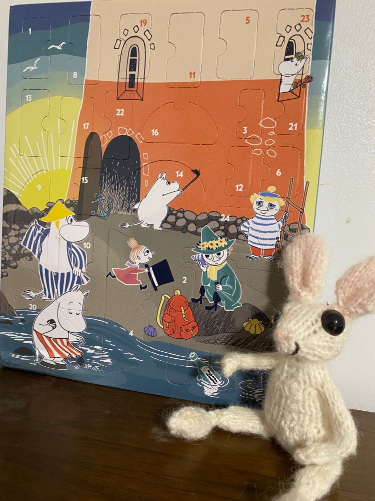 A small knitted white rabbit sits beside a Moomin advent calendar with a seaside scene of a lighthouse and a rocky beach.