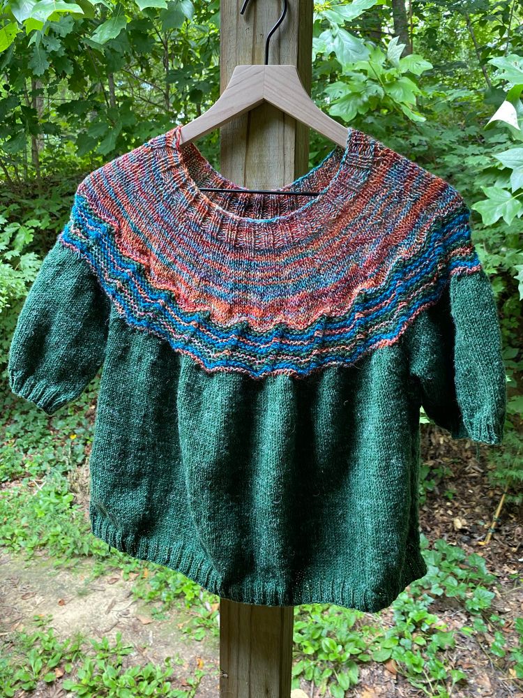 A short sleeved handknitted sweater hangs out on the back porch. It has a multicolored yoke, blue and green zigzag accents and a tweedy forest green main body and sleeves. It was a delight to knit.