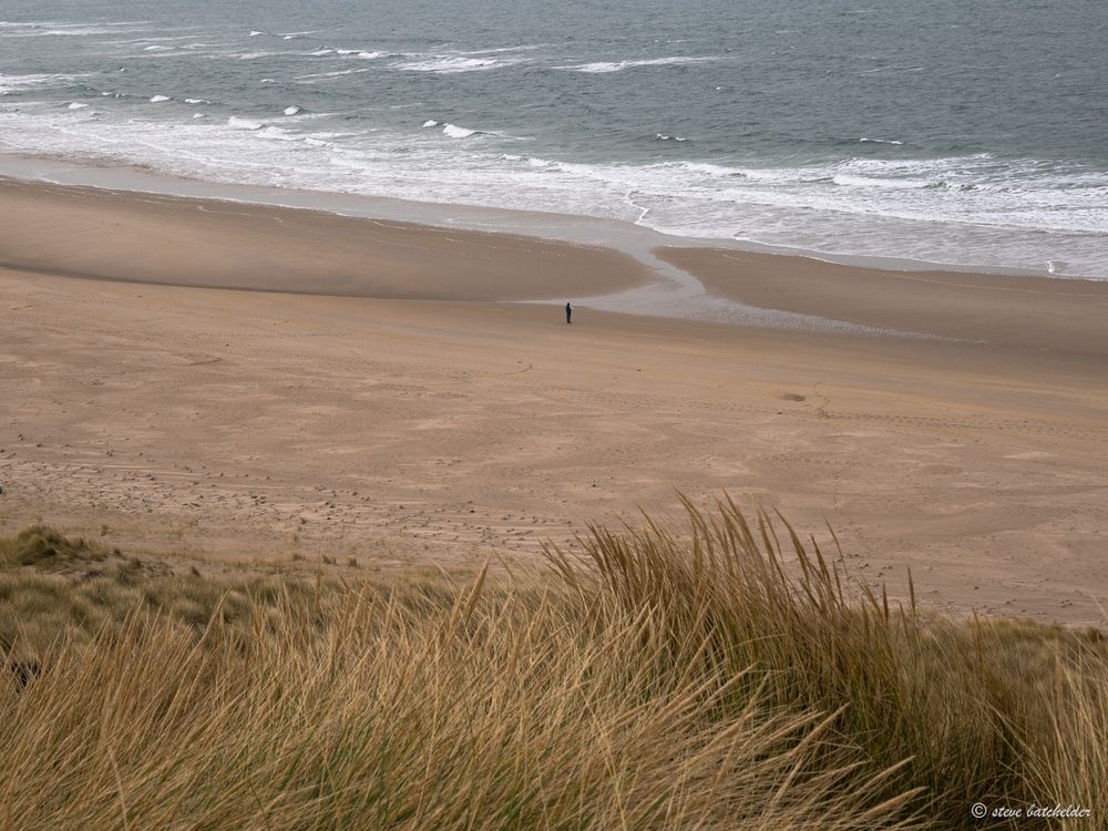 Image shows a sandy beach with the distant figure of a person in dark clothing staring out to sea. Marram grass is visible in the foreground followed by a wide stretch of sand leading down to the sea where the breaking waves create a line of white wth the sea behind it at the top of the image.