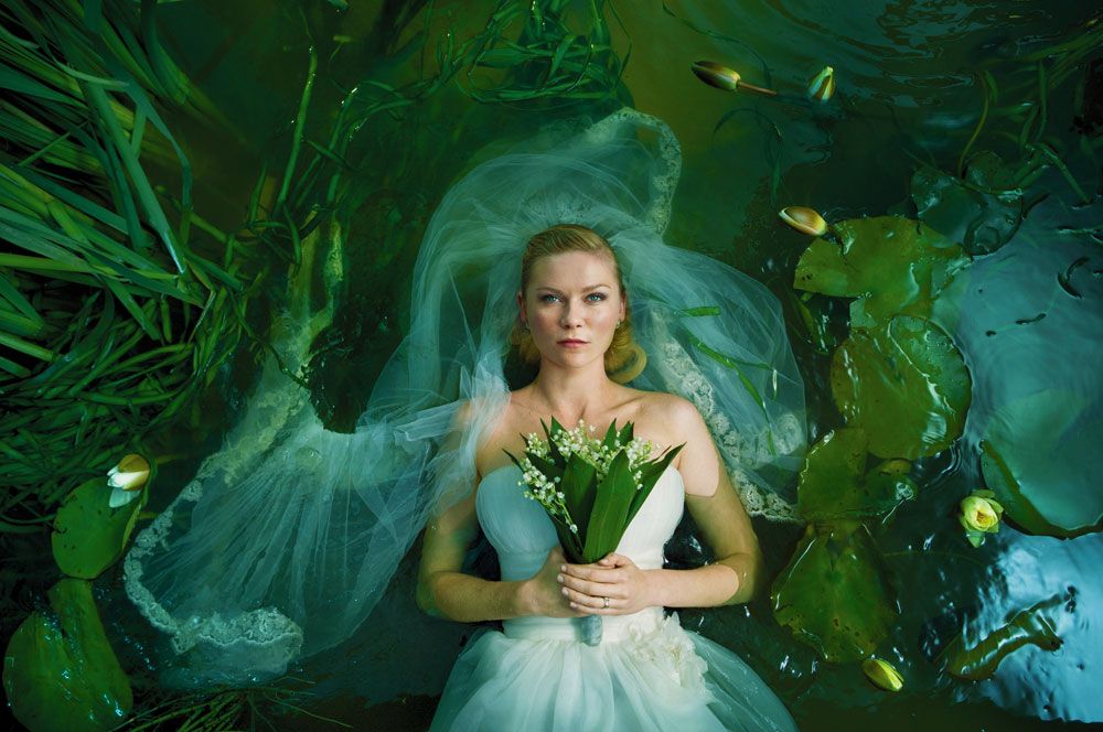 Image from the movie Melancholia with a blond woman in a bridal dress floating in a pond amongst lily pads, staring upwards, clasping a bouquet to her chest, veil swirling. Reminiscent of Ophelia.