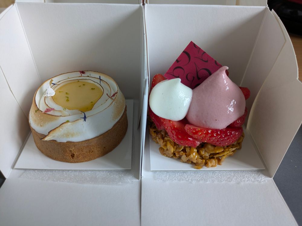 Photo de deux desserts individuels dans leurs boites en carton : à gauche une jolie tarte citron meringuée avec des fleurs décoratives sur la meringue, à droite un genre de fraisier avec un croustillant corn flakes, des quartiers de fraise, et de la mousse sur le dessus