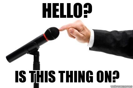 Hand tapping a microphone on a mic stand. The person is wearing a suit jacket. Overlaid text reads "HELLO? IS THIS THING ON?"