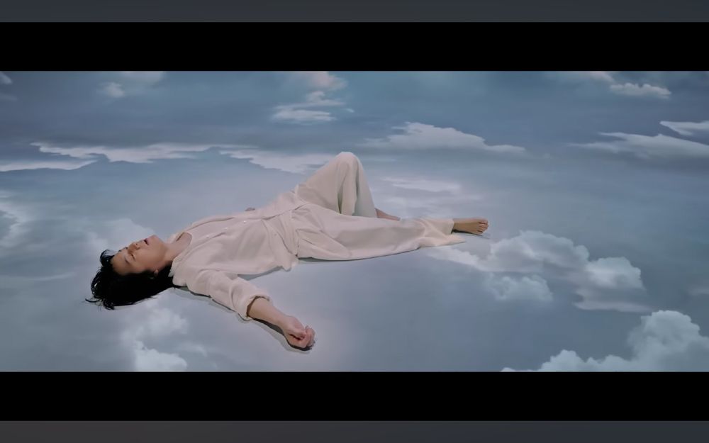 Screenshot from the music video for Fukuyama Masaharu's Soubou, showing him dressed in white and lying on clouds in a blue sky.