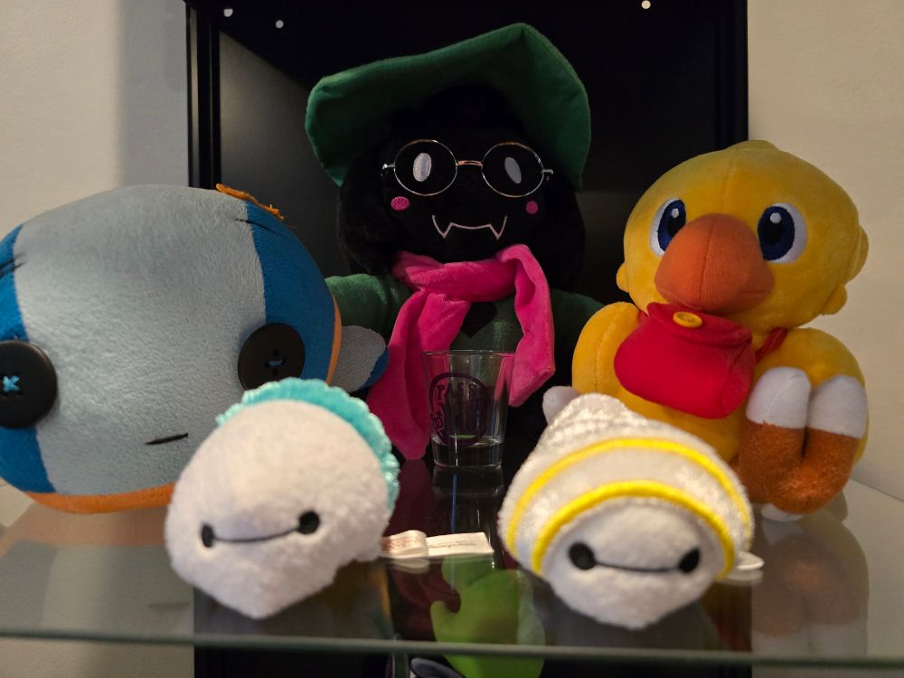 A picture of a glass shelf holding several plush figures; the "Scumbag" from the game Bastion, two plushies in the shape of sushi with the face of Baymax, a Chocobo courier, and Ralsei from Deltarune. A shot glass is obscured in the center of the photo. It has the phrase "Pass the Yaoi" printed on it.