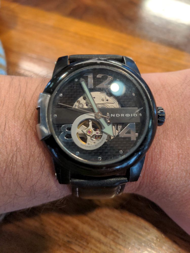 A white man's wrist adorned with a black mechanical watch branded android. 