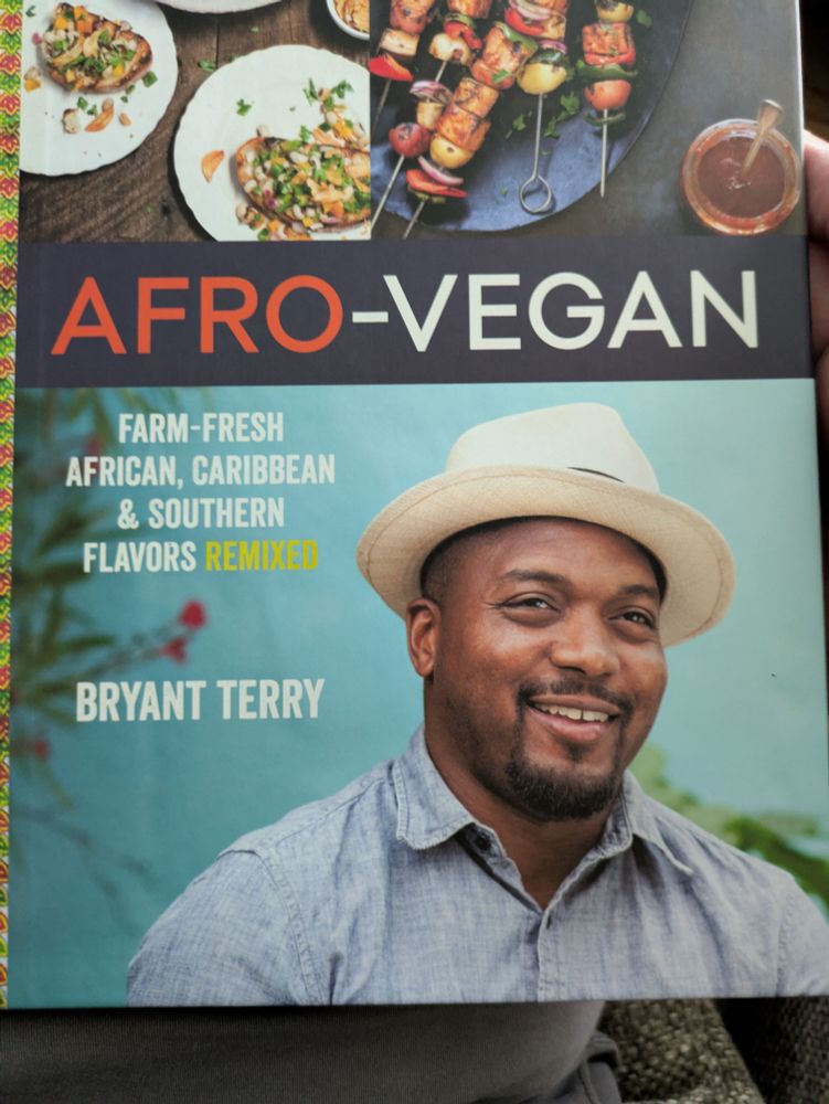The cover of a cookbook titled "Afro-vegan" on the cover are two dishes, Texas caviar & veggie kebobs, above a picture of the author, a dapper black man in a white hat and light blue shirt. His name is Bryant Terry. 