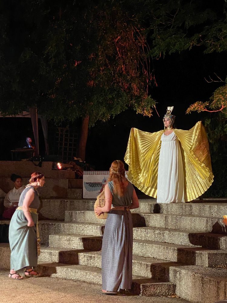Actress playing Cleopatra holding out her arms with large golden cloak flowing. Her two ladies in waiting are at the bottom of the steps and she’s at the top. Outdoor theatre in the twilight.