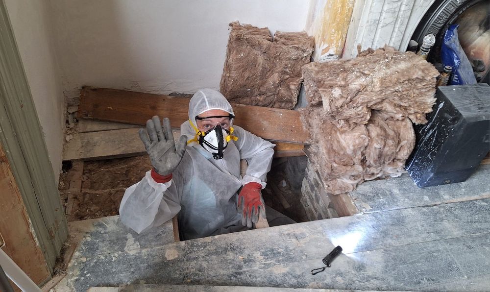 Kate halfway through floorboards and into cavity beneath. Waving, wearing a full white dust suit, mask, goggles and gloves. 
