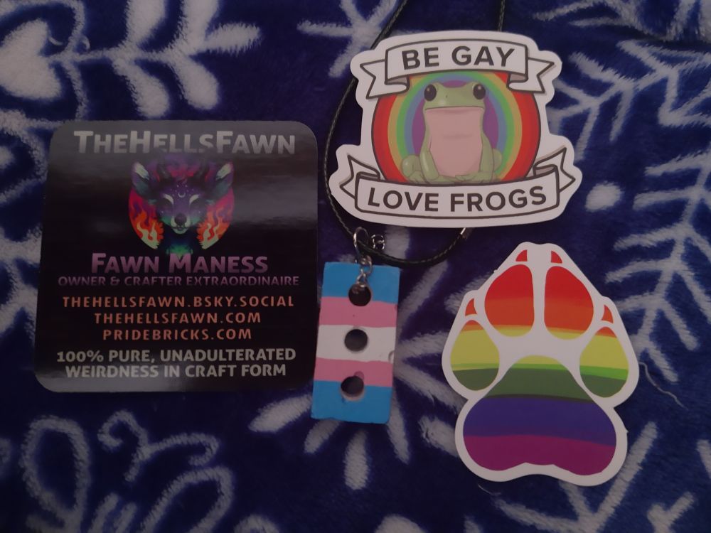 A necklace of a brick painted the colors of the trans pride flag, as well as a sticker of a paw colored like the gay pride flag, and a gay pride sticker about appreciating frogs.

All of this was sent to me by @thehellsfawn, who has several websites including pridebricks.com and thehellsfawn.com