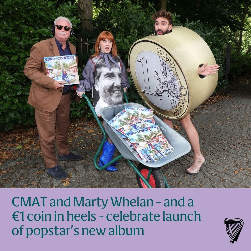 A photoshoot pic taken in St Stephen's Green park with CMAT wearing a dress with Daniel O'Donnell's face on it and pushing a wheelbarrow full of copies of her new album. Marty Whelan stands beside her holding a copy of the album, wearing headphones. And her bandmate Púca stands on the other side, wearing a giant one euro coin costume and high heels. The caption reads: CMAT and Marty Whelan – and a €1 coin in heels – celebrate launch of popstar’s new album