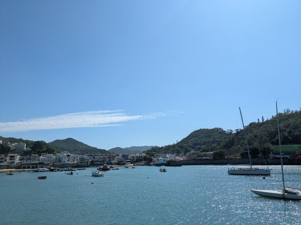 A photo of a blue sky with one streaky cloud on the left. There are boats on the water and mountains in the background. The photo was taken on Lamma Island in Hong Kong.
