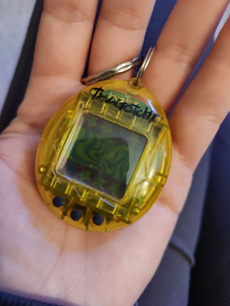A photo of a hand holding a yellow Tamagotchi digital "pet" simulator circa the late 1900's. 