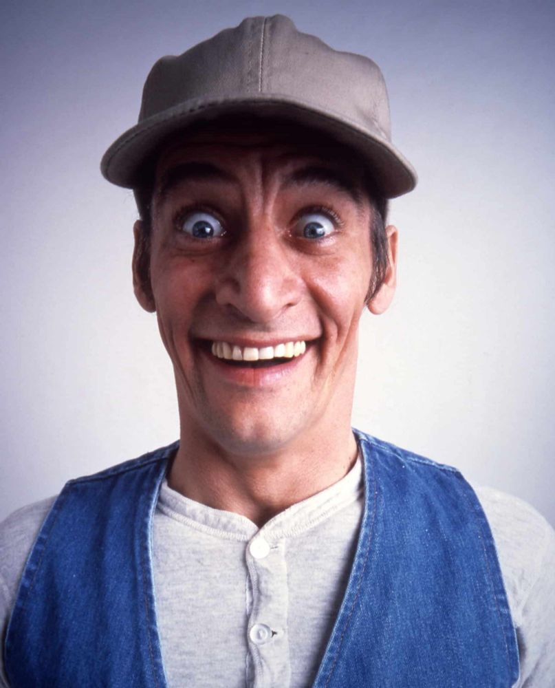 A headshot (mugshot, perhaps) of TV character Ernest P Worrell cracking a concerningly toothy smile. He's a middle-aged white man wearing a tan-gray ball cap, a denim vest and a white Henley shirt. 