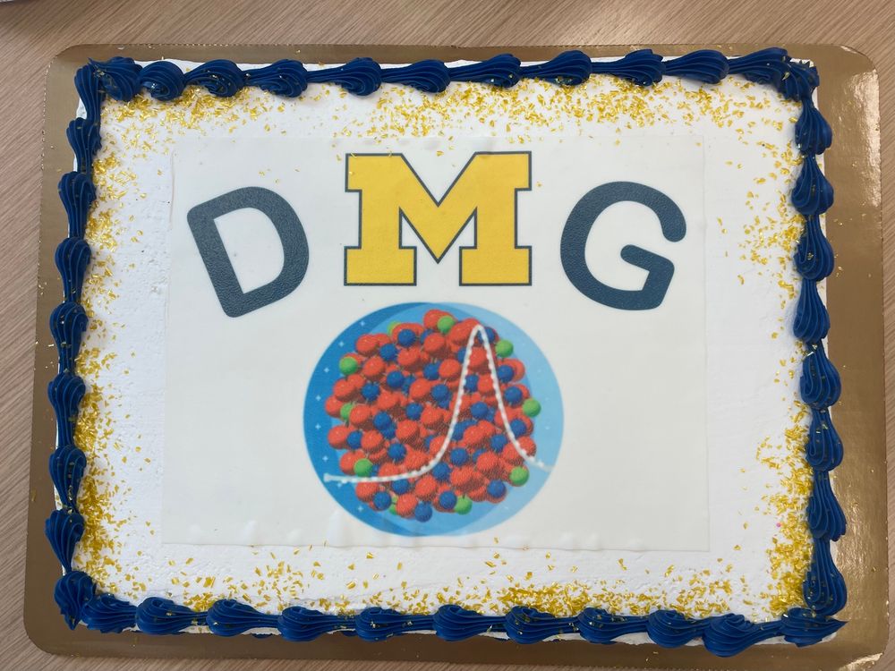 A cake with blue and maize colored decoration, printed with DMG, the M resembling the University of Michigan logo and a scientific visualization of a localized surface plasmon resonance in a doped nanocrystal. 