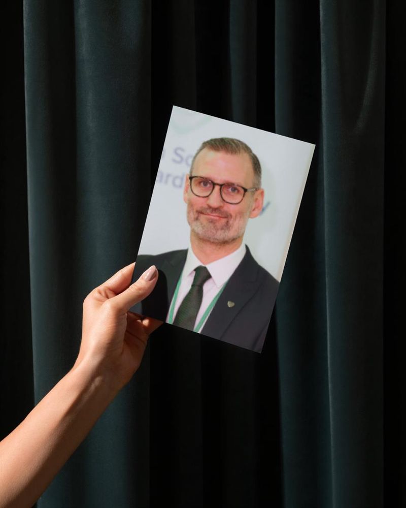 A picture of a hand holding a photo of Shaun against a green curtain