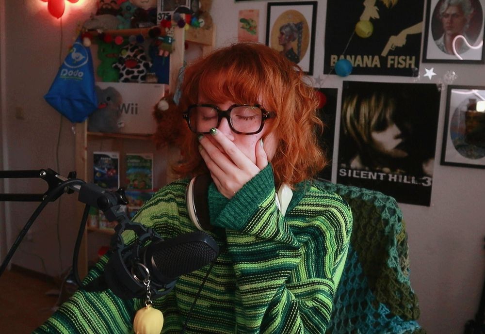 emmy in her room, wearing a green stripey jumper and has her hand to her face with her eyes closed because she is very tired