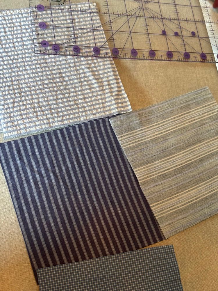 Squares of a variety of blue striped and checked fabrics, cut for quilting and spread out, sitting on a grey ironing board with a clear quilting ruler at the top right of the frame. 