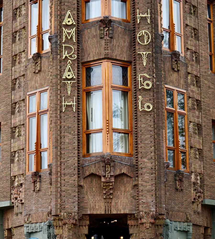 Pan coupé à l’angle d’une façade en briques chargée de petites sculptures et de motifs composés de briques en relief. Des lettres imitant un style rétro indiquent à la verticale “Amrath Hotel”. L’impression générale est foisonnante. 
