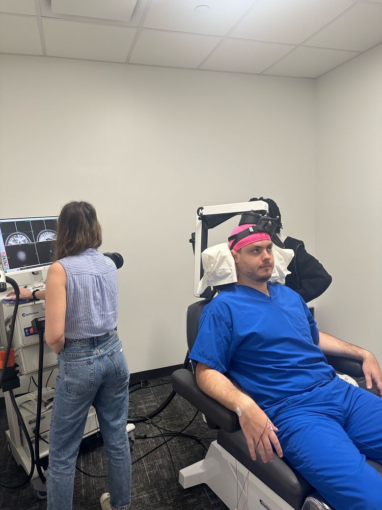 An adult male in blue scrubs and a pink swim cap receives MRI guided TMS to the motor cortex. A female stands in the background looking at the MRI images 