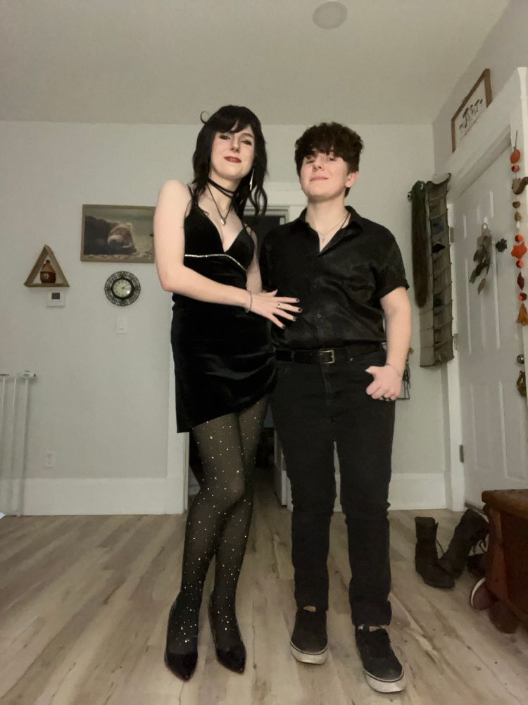A butch femme couple standing next to each other wearing all black with little sparkly embellishments