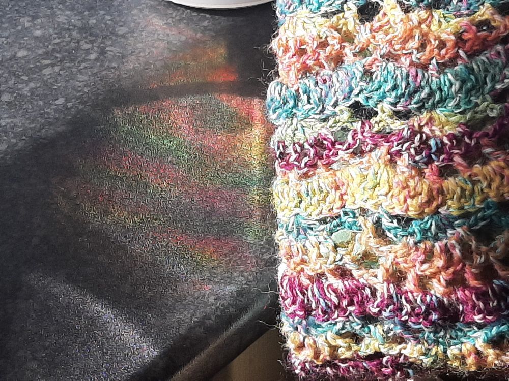 Rainbow of light caused by a prism reflected onto a dark counter top, fan lace crochet in similar rainbow like colours held next to the rainbow of light for comparison. 