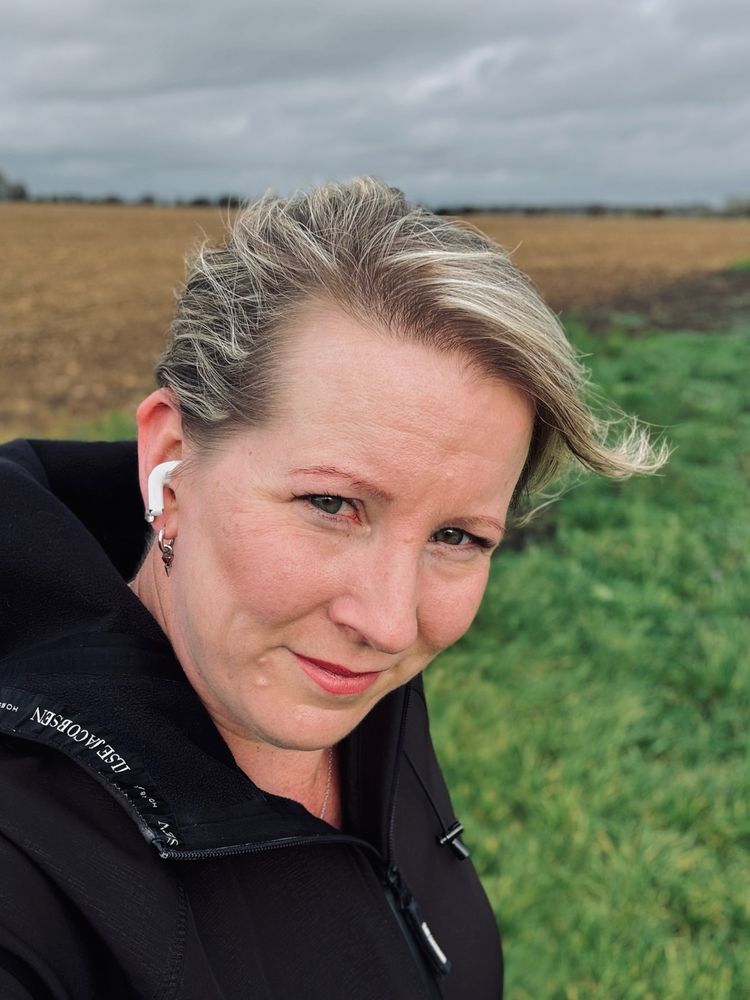 A selfie of me, a middle-aged, fair-skinned woman wearing a black coat, with short blonde hair blowing all over the place. 