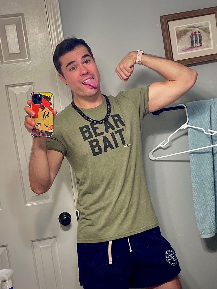 Man with his tongue out wearing green shirt that says BEAR BAIT while flexing into camera