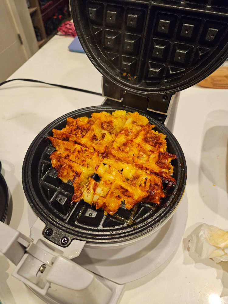 a waffle maker holding a waffle-shaped amalgamation that's mostly orange with grill marks. At least one intact cavatappi is visible
