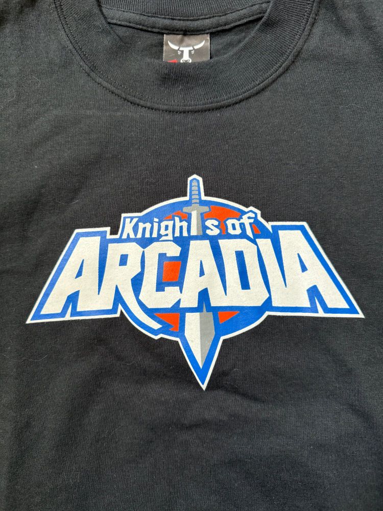 A black T-shirt with the logo for "Knights of Arcadia," consisting of the words "Knights of" centered above the word "Arcadia" in larger text, with a sword pointed down cutting through both. The logo incorporates the orange and blue of the Penny Arcade brand.