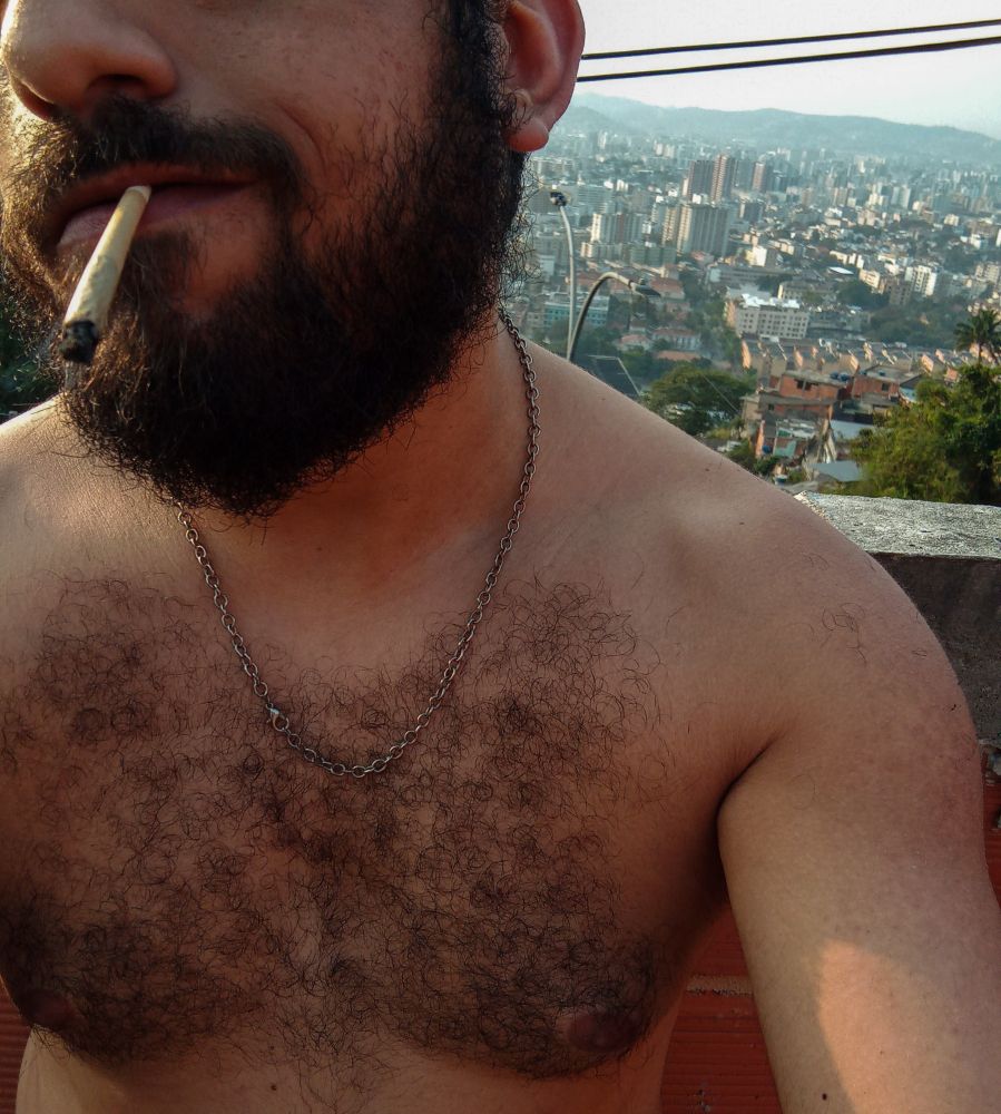 Foto de um homem pardo sem camisa com uma correntinha como colar e um cigarro na boca.
Ao fundo pode se ver de cima parte da cidade.