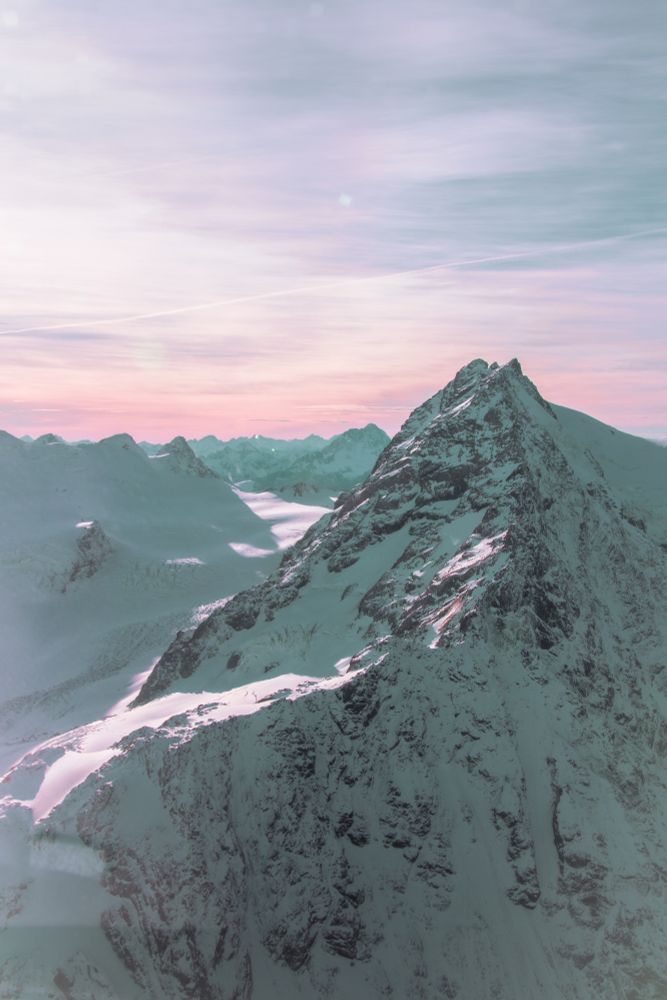 The picture shows the snow-covered mountain peaks of New Zealand's Southern Alps. It is taken mid-flight from a helicopter. As such, the viewer is at eye level with the towering mountain peak that forms the foreground of the picture. The approaching sunrise tints the sky a soft orange and pink, giving the scene a serene and untouched beauty.