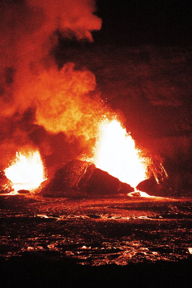 Volcanic eruption of Kilauea on Hawaii's Big Island. The picture is taken at night, while the erupting lava illuminates the sky. Great plumes of smoke trail from the volcano, while a glowing trail of lava snakes along the ground. All along the caldera, the ground is glowing, and lava is showing through a million tiny cracks.
