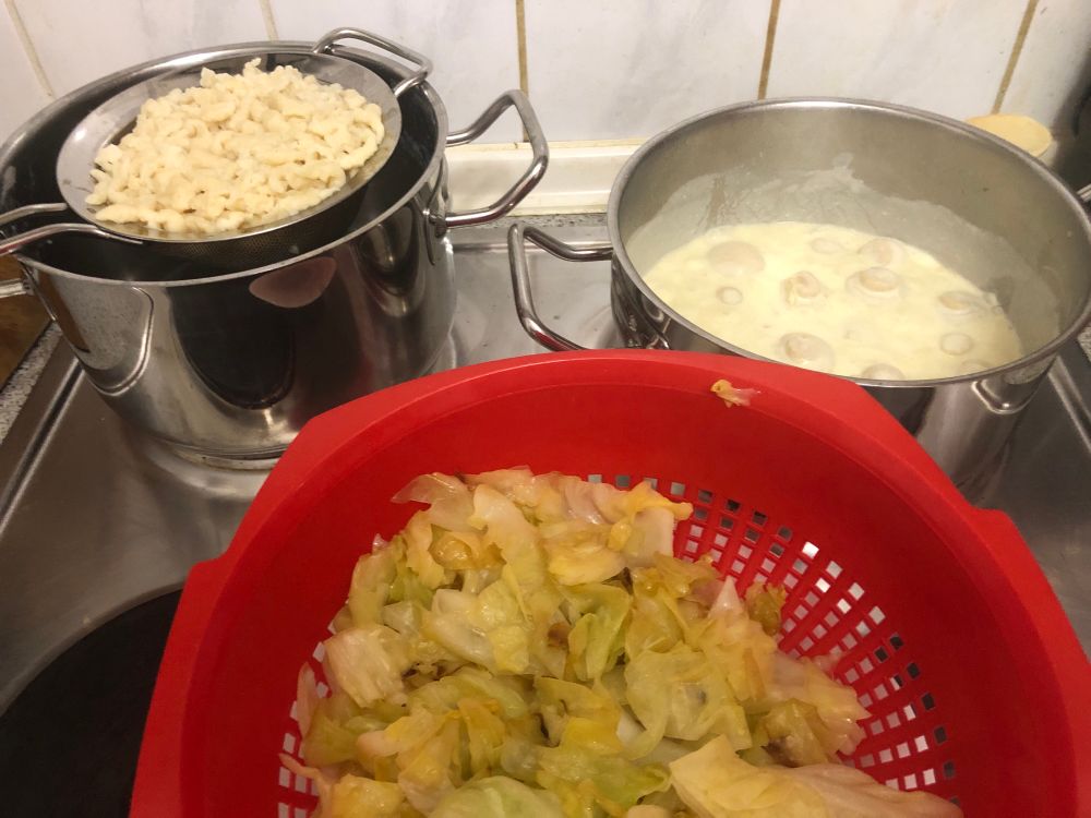 Auf dem Herd hinten links selbstgemachte Spätzle in einem Edelstahlsieb über einem Edelstahltopf, rechts daneben eine helle Sauce in der kleine braune Champignons schwimmen. Davor in einem Plastesieb angeschmorter geschnittener Spitzkohl. 