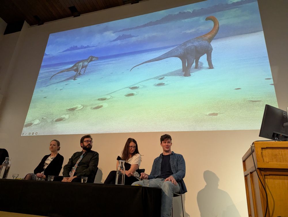 Four palaeontologists in a Q&A session in front of some hack's painting of the Dewars Farm Quarry dinosaur tracks, as it might have appeared in the Jurassic.