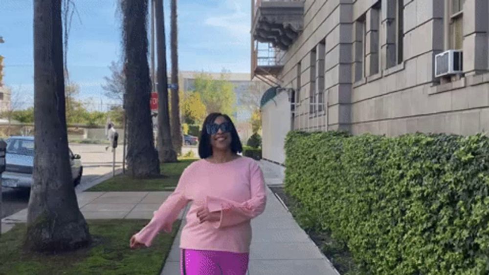 a woman wearing sunglasses and a pink sweater is walking down the sidewalk