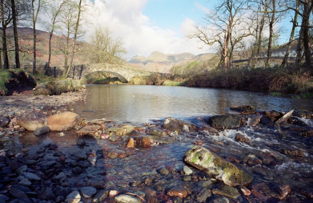 Colour photo (taken on Kodak Ektar film) of Gt Langdale Beck where it flows over a small barrier of rocks, just below a stone bridge, with the Langdale Pikes in the background