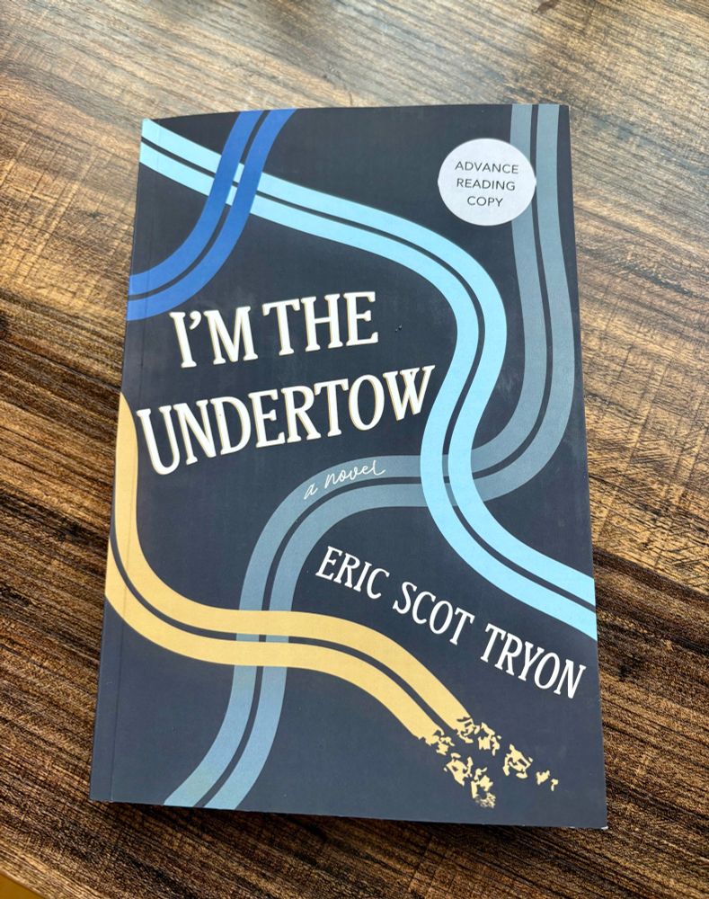 Book on a brown table. The book is dark blue with various curvy lines (shades of blue and beige) weaving throughout. The title in the middle reads I’M THE UNDERTOW. The author’s name at an angle in the bottom right corner reads Eric Scot Tryon. 