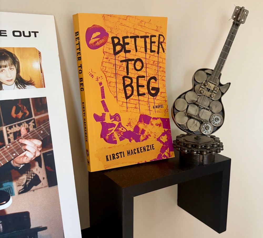 Centered in the photo is an orange and magenta book  on a shelf called BETTER TO BEG by Kirsti MacKenzie. To the right of the book  is a small metal sculpture of a guitar. To the left of the book is a partial  peek at an album cover. 