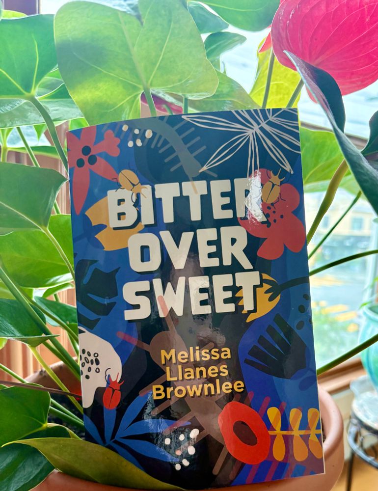 A book (BITTER OVER SWEET by Melissa Llanes Brownlee) sitting amongst a plant with big green leaves and a red flower. 