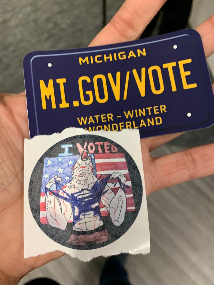 Photo of a hand holding a small blue and gold Michigan license plate shaped magnet that says “MI.GOV/VOTE” and a round handrawn sticker that says I voted in blue and red and features a werewolf standing in front of the American flag ripping its shirt apart and howling presumably with pride for having fulfilled their civic duty. 