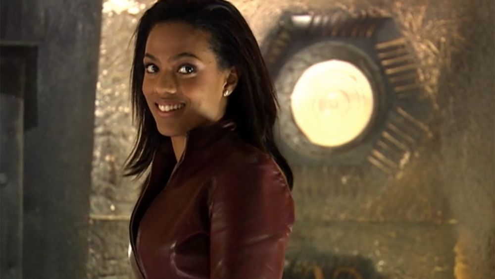 Photo of Martha Jones a companion of Doctor Who. A young pretty Black woman smiling  with her straightened, shoulder-length, dark hair down wearing a burgundy jacket and standing next to a round yellow light on the TARDIS. 