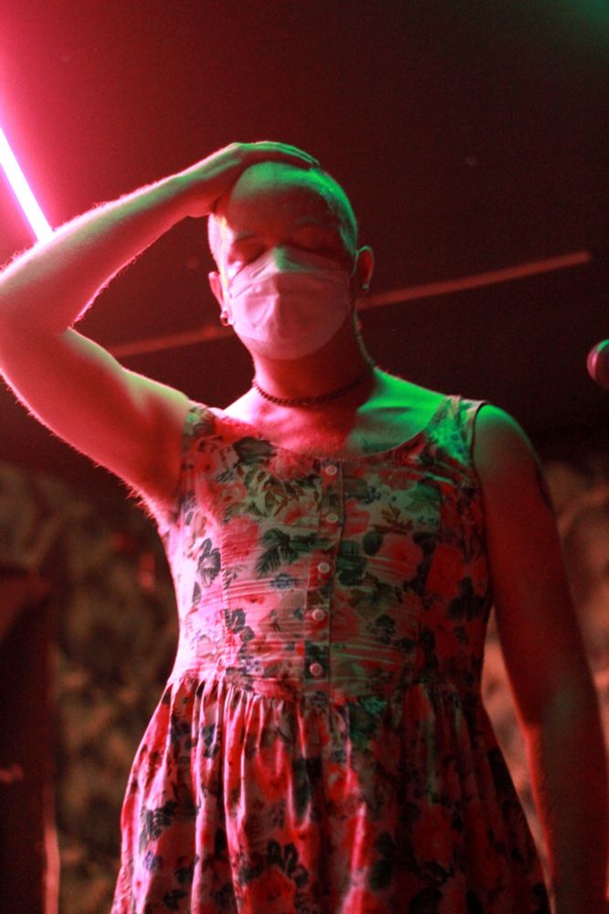 A photo of a person illuminated by greens and reds. she's wearing a floral dress and white mask