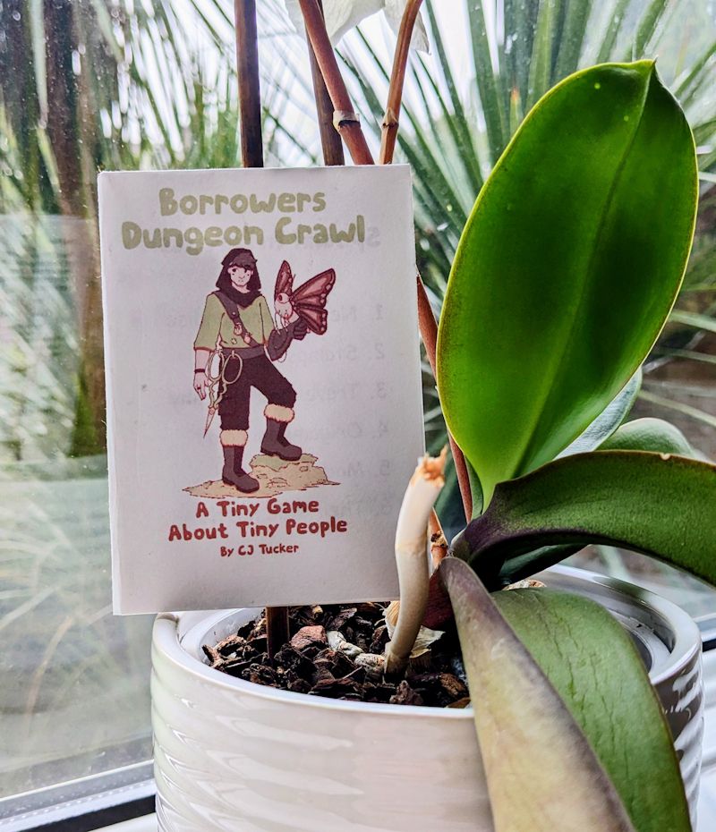 The game Borrowers Dungeon Crawl placed inside a plant pot. 

On the cover, beneath the title, is an illustration of a light-skinned person. They have short hair and a hood, on one hand rests a large moth, and on their belt is a large pair of embroidery scissors. Beneath the illustration is the subtitle: "A Tiny Game About Tiny People by CJ Tucker"