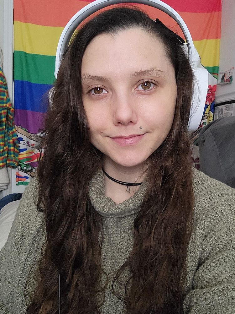 a rizzi selfie- a woman with long brown hair wearing a white headset, black choker necklace and sage green sweater