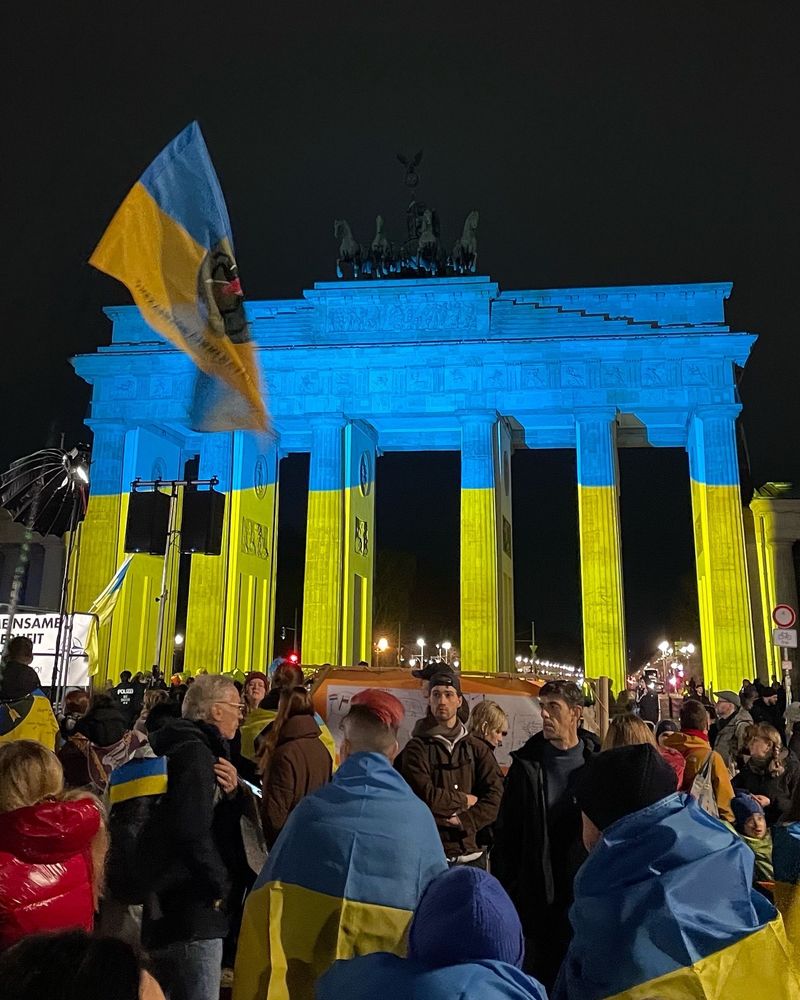Foto von der Demo in Berlin am 24.2.2025, dem Jahrestag der russischen Vollinvasion in der Ukraine. Zu sehen sind Menschen gehüllt in Ukraine-Flaggen, im Hintergrund das in den ukrainischen Farben Blau und Gelb leuchtende Brandenburger Tor. 
