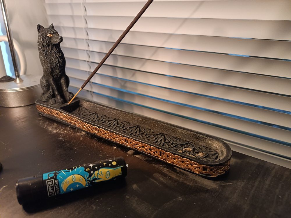Black incense holder with a small sculpture of a wolf, and vaguely norse style knotwork sculpted into the sides. The wolf's eyes are painted gold, as is the knotwork.