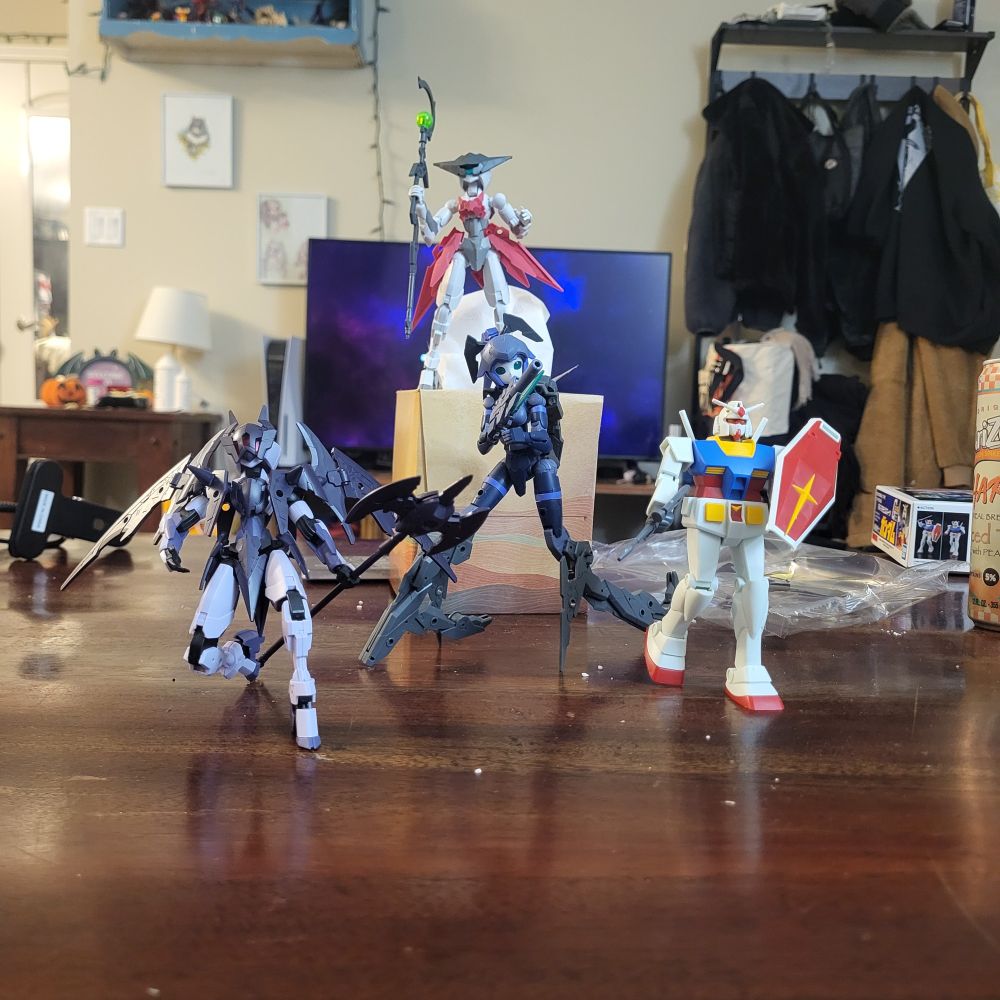It is a picture of four gun pla in a living room. The background is a common front wall of a livingn room with a jacket rack, TV and side table. The four gun pla are arranged from left to right: purple cat girl mech with wings, blue cat girl mech with Grey attachments, the Rx-78-2 gundam, up on top of a tissue box is a white and red cat girl mech wielding a staff with a green orb 