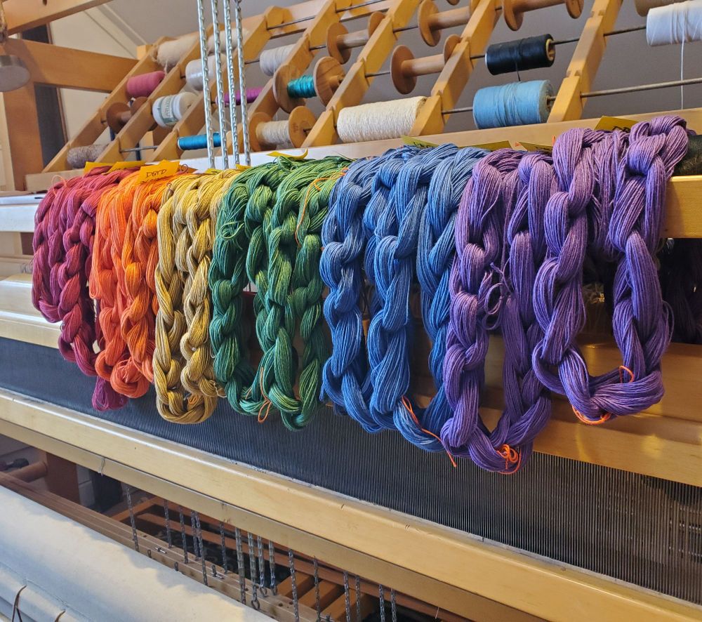 A rainbow of bundles of chained string draped over the hanging shelf of a Cranbrook loom. Viewer can see a peek of the front beam and apron in fore, treadle chains, reed and beater, and a spool rack of various yarns in the background.