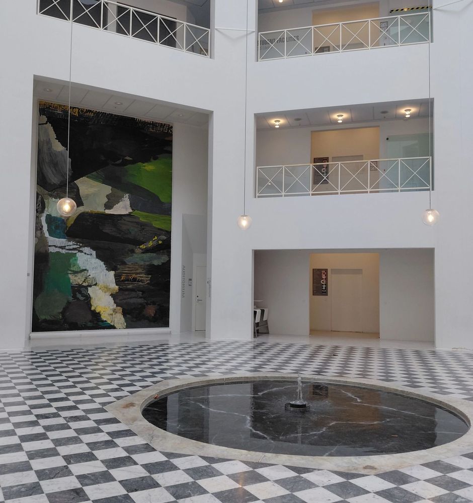 A building lobby, white shades, black-and-white tiled floor, a fountain in the middle, and an abstract greenish painting in the background.