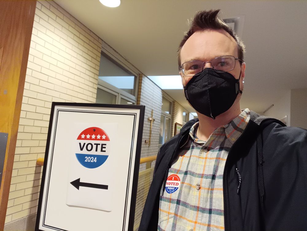 A man wearing a black face mask and glasses, sporting an "I Voted Today" sticker on his shirt, and standing next to a sign that reads, "VOTE 2024"